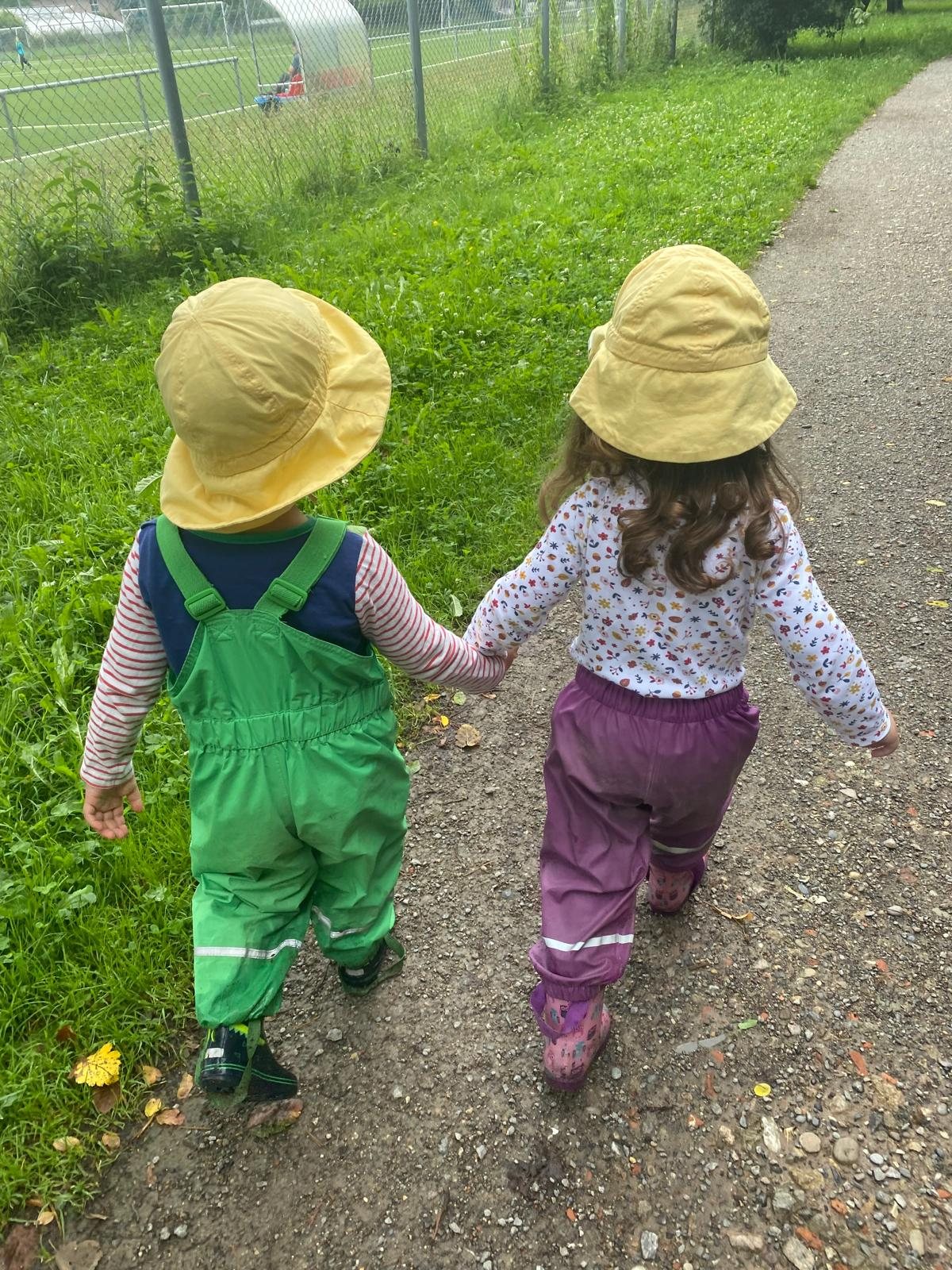 Zwei Kinder in Matschhosen und Sonnenhüten gehen Hand in Hand einen Feldweg entlang