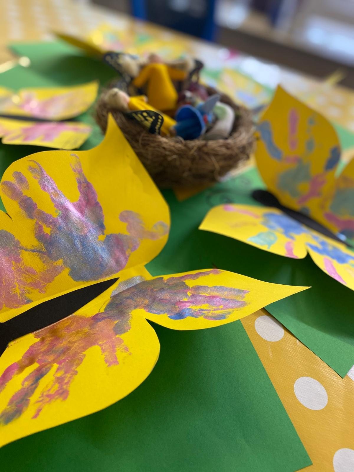 Gelbe Papierschmetterlinge mit bunten Handabdrücken, im Hintergrund ein Vogelnest mit Blütenfeen